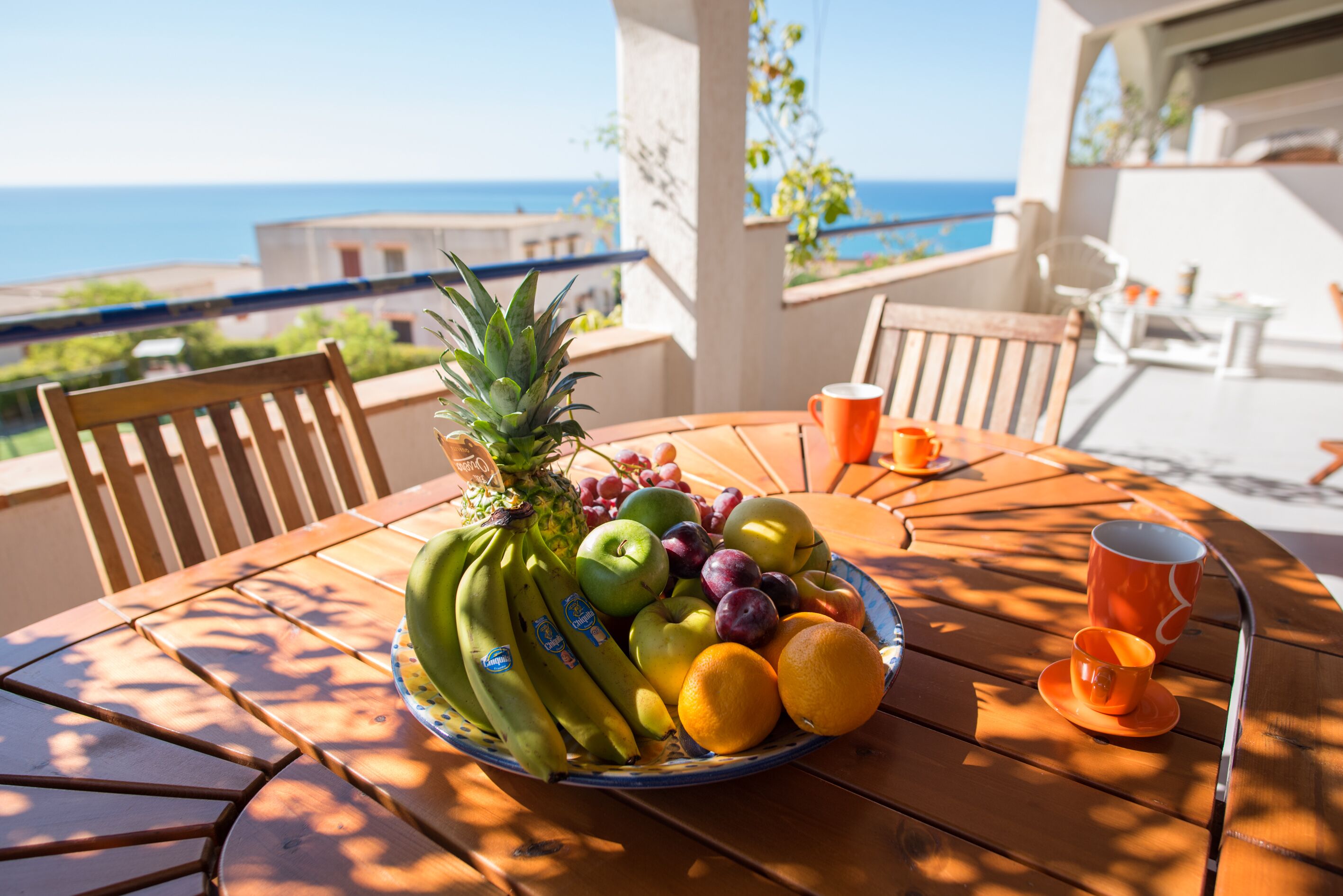 A round wooden table with a fruit bowl containing bananas, apples, grapes, and oranges, and two orange cups on a balcony overlooking the sea.