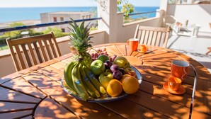 Un tavolo rotondo in legno con una ciotola di frutta contenente banane, mele, uva e arance, e due bicchieri arancioni su un balcone con vista sul mare.