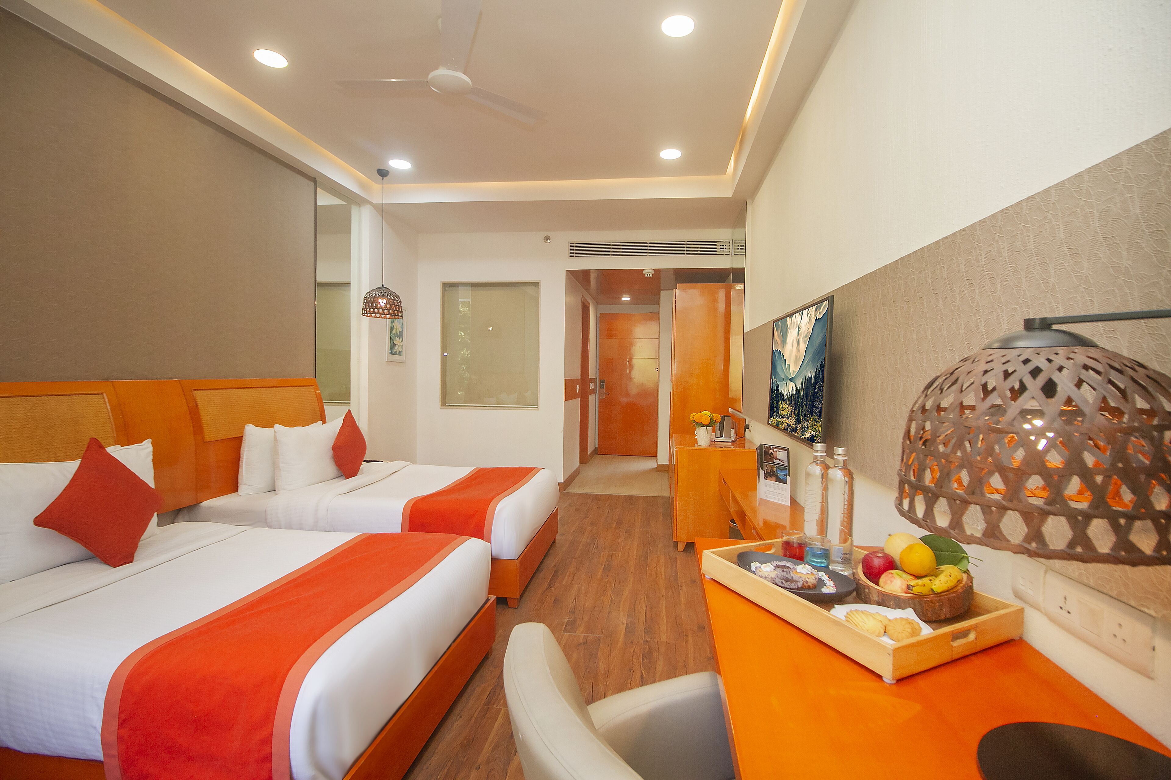 A hotel room with two beds, a desk with a fruit tray, and a decorative lamp.