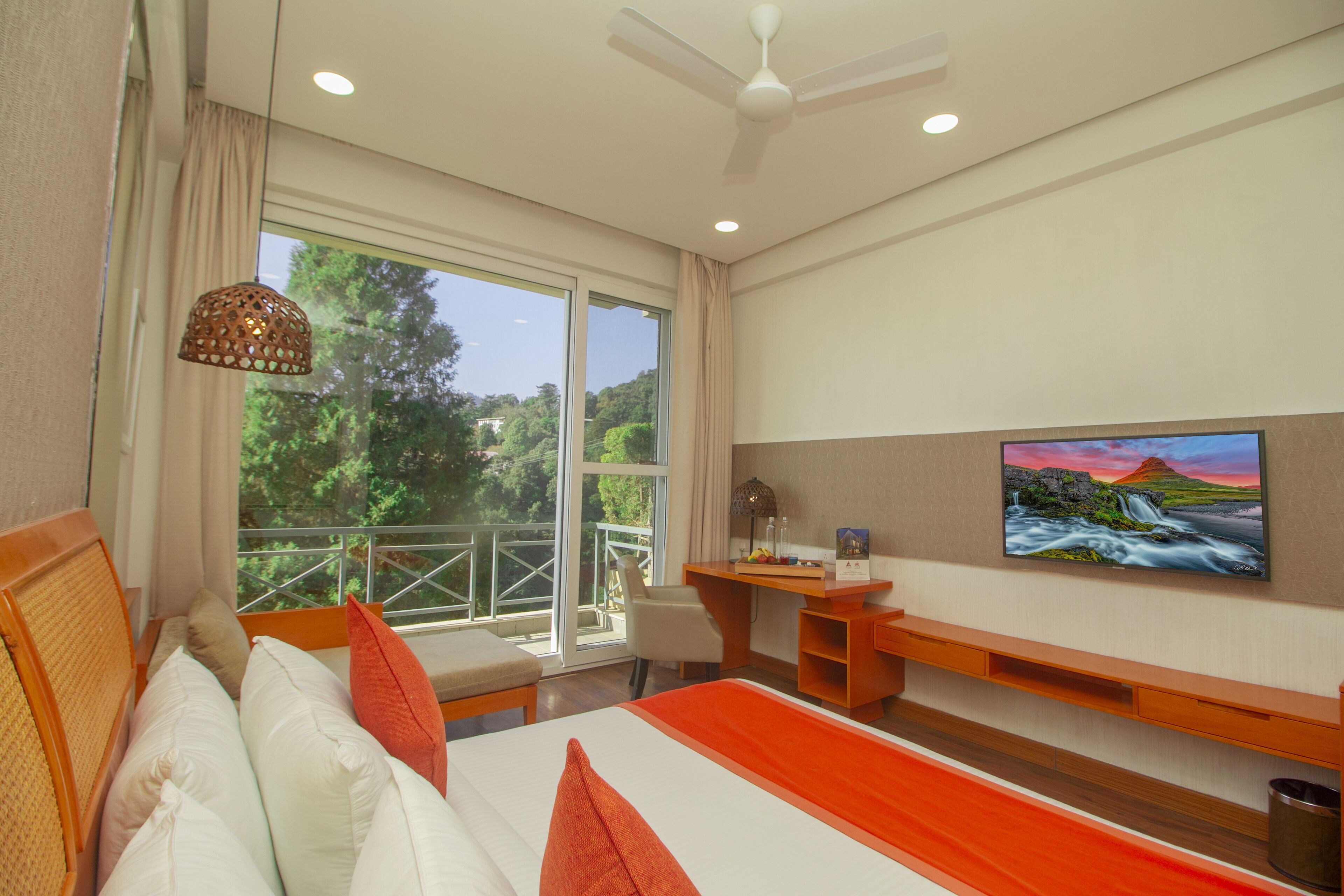 A hotel room with a bed, a desk, a chair, a TV, and a balcony with a view of trees.