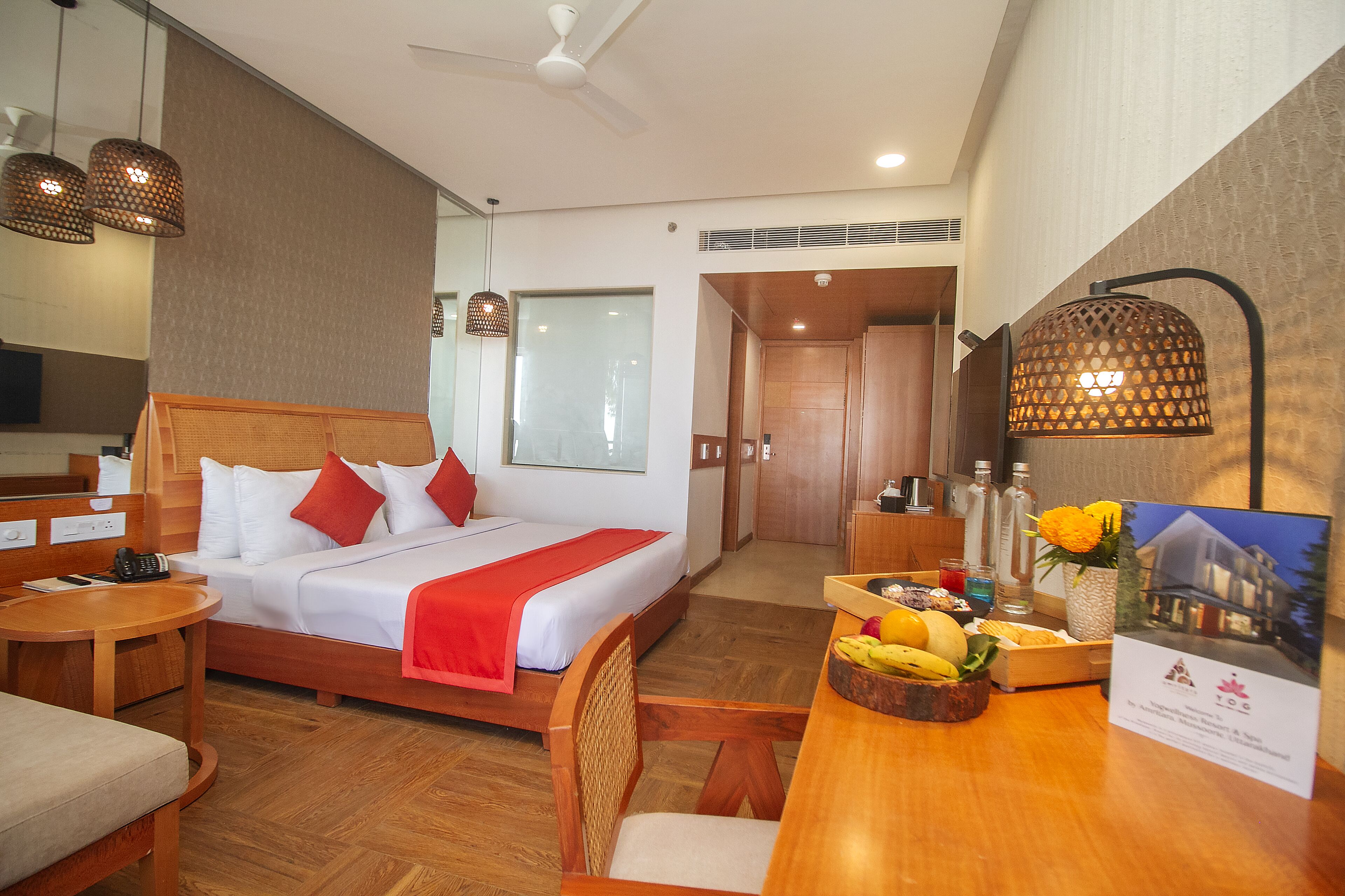 A hotel room with a bed, bedside table, chair, and a desk with a fruit bowl and a brochure.