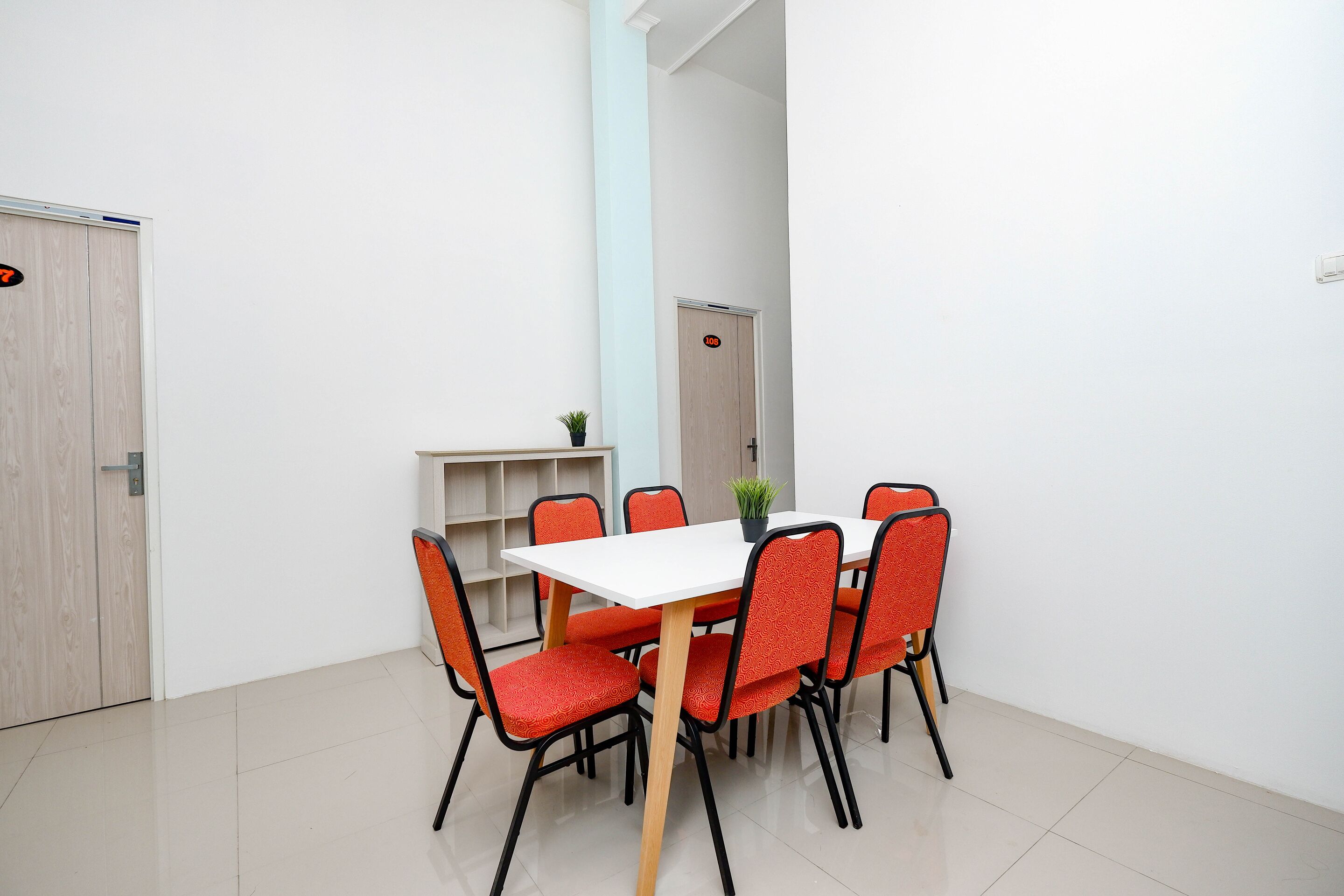 A dining area with a white table and orange chairs, a small plant on the table, and a light blue pillar.