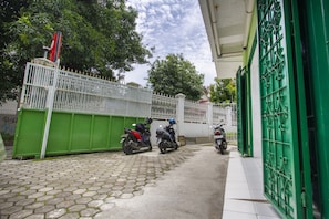 Un chemin pavé, bordé d’une clôture verte et surmonté d’un portail blanc, mène à un bâtiment doté d’une porte verte.