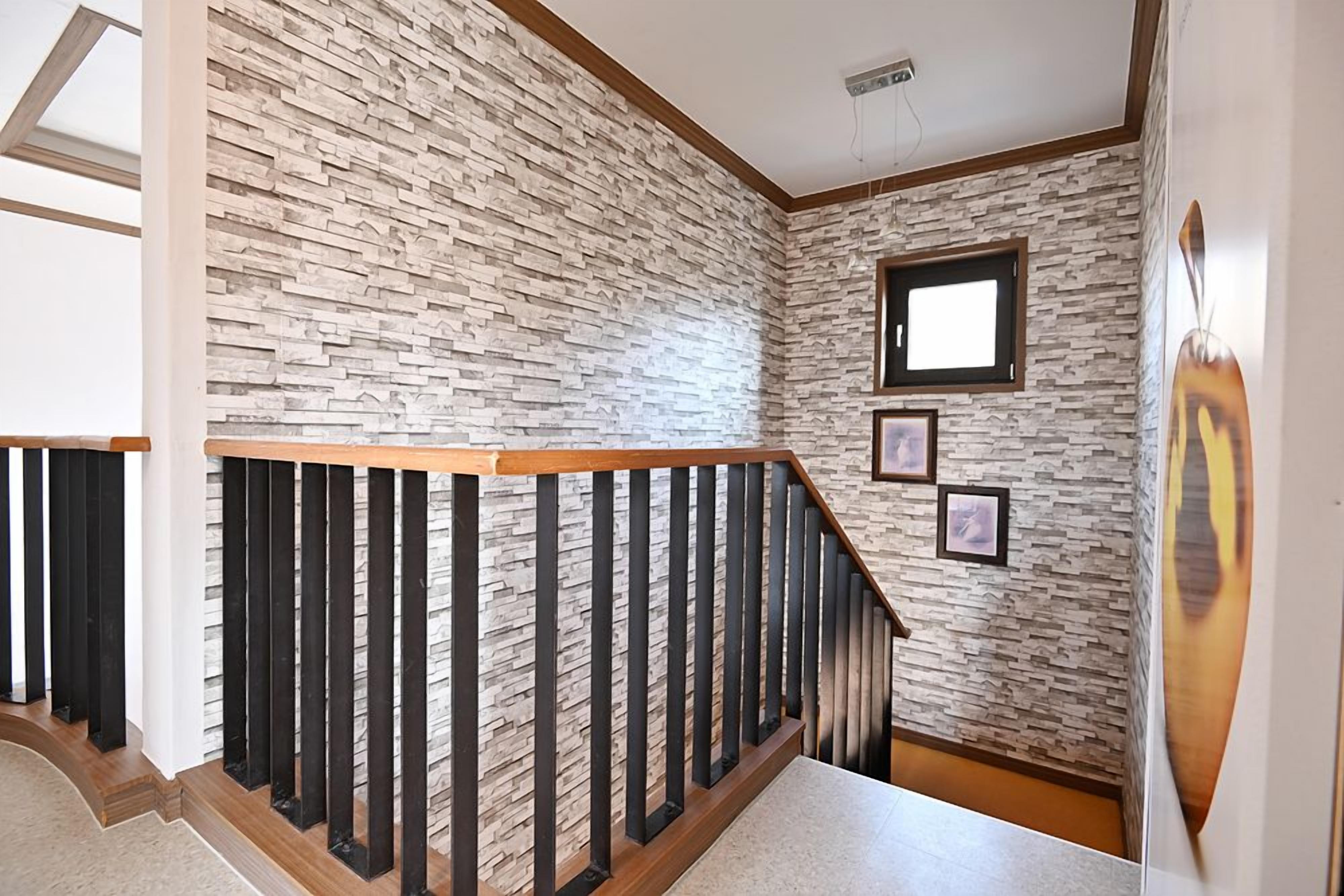 A staircase with a wooden handrail and black balusters, leading to a room with a stone wall and a small window.