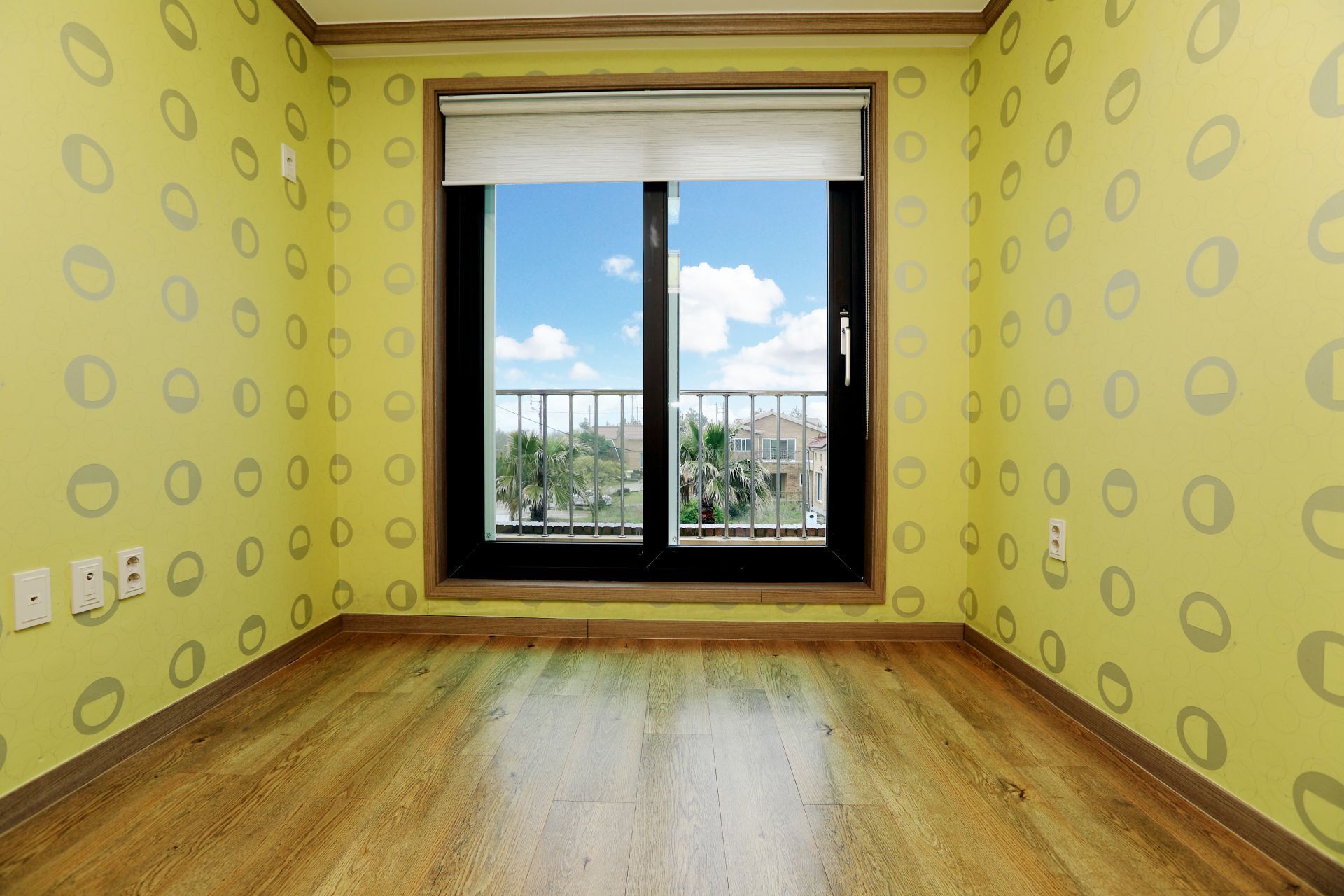 Una habitación con paredes amarillas, un ventanal grande con persiana blanca y suelo de madera.