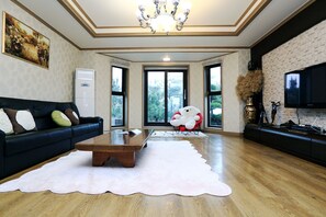 A spacious living room with a dark sofa, a white coffee table, and a large window offering a view of trees. - Ocean Sky Village Pension (Jeju City)