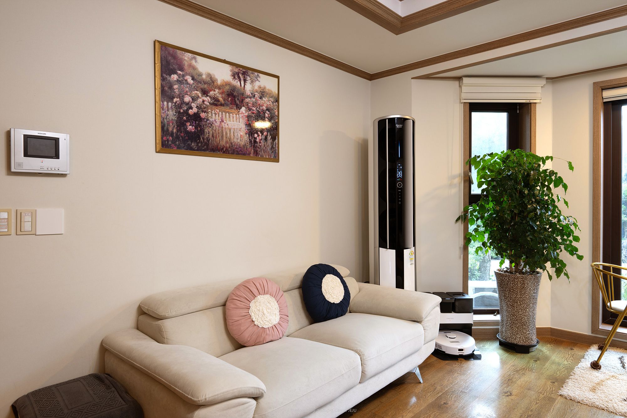Un soggiorno con un divano beige, un quadro, un elettrodomestico alto, una pianta in vaso e una finestra.