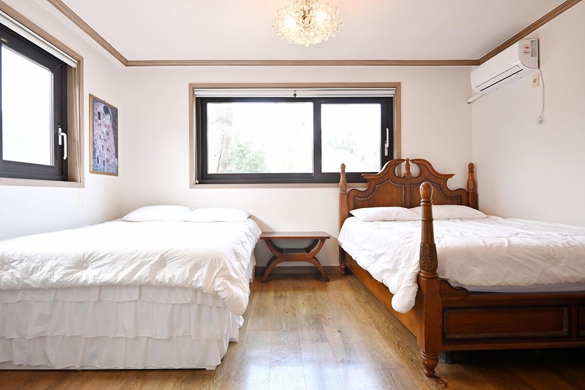 A bedroom with two single beds, a small table, a window with blinds, and an air conditioning unit on the wall.