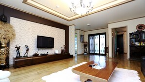 Un salon spacieux avec une table basse en bois, un téléviseur à écran plat fixé sur un mur en briques blanches et un lustre.