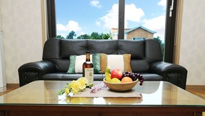 Un salon avec un canapé en cuir noir, une table basse en verre sur laquelle se trouvent un panier de fruits et une bouteille, et une vue sur une maison à travers de grandes fenêtres.