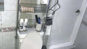 Un baño con inodoro, zona de ducha, secador de pelo y estante con artículos de aseo.