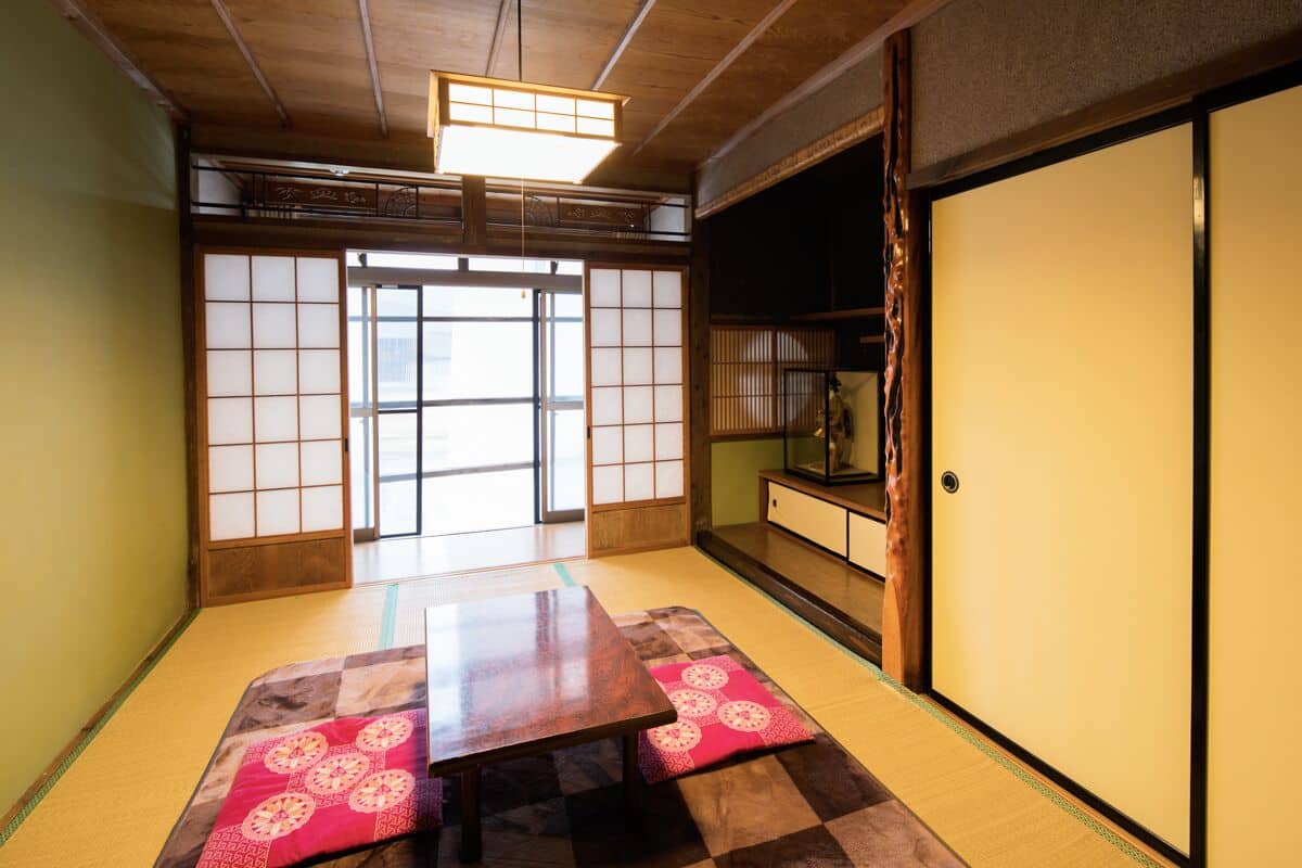 Una habitación japonesa tradicional con suelo de tatami, una mesa de madera baja, puertas correderas y vistas a través de un biombó shoji.