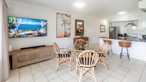 A modern living room with a large TV, a dining table with chairs, a kitchen area, and a view of the ocean.
