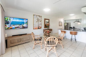 A modern living room with a large TV, a dining table with chairs, a kitchen area, and a view of the ocean.