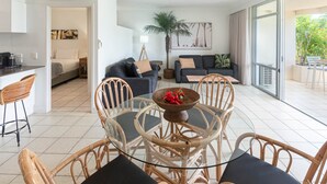 A modern living room with a glass dining table surrounded by wicker chairs, a sofa with cushions, a potted palm tree, and a balcony with a view of greenery.