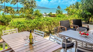 An outdoor dining area with wooden tables and chairs, overlooking a beach and palm trees.