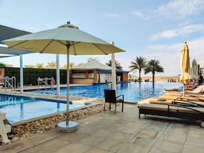 A swimming pool with lounge chairs and umbrellas, surrounded by palm trees and a modern building.