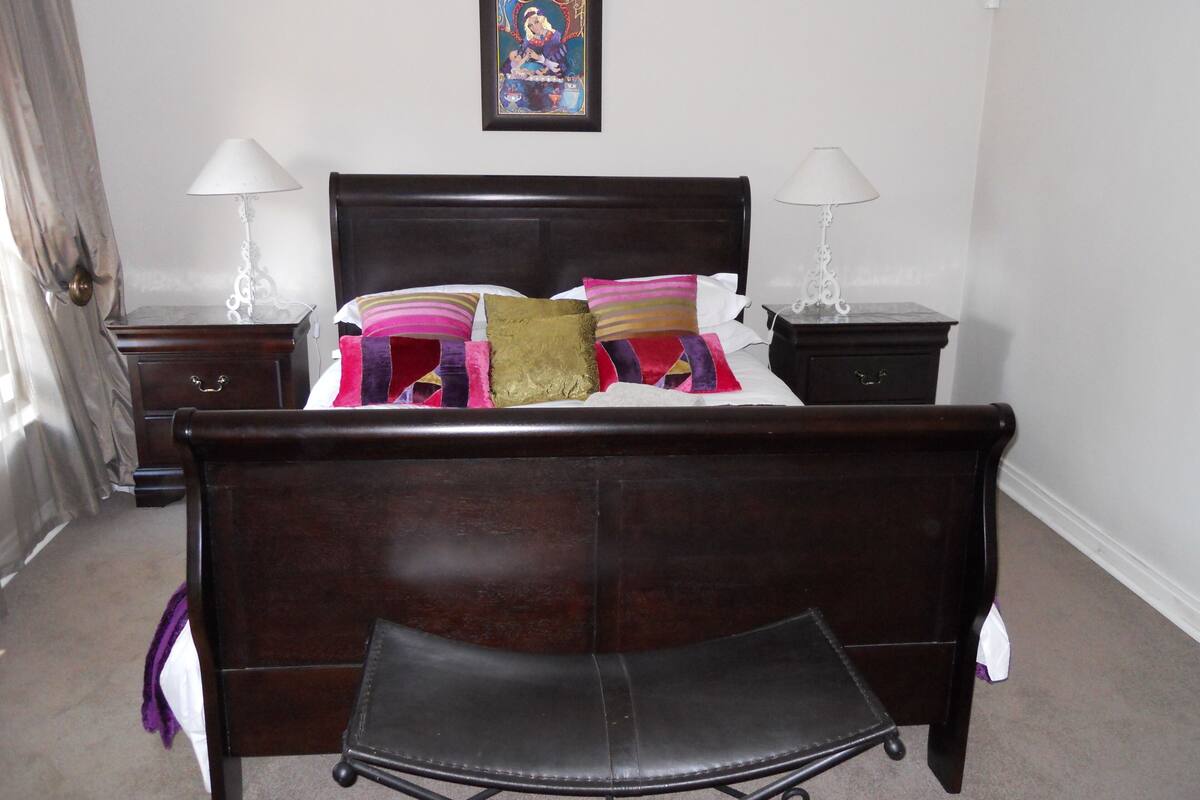Une chambre avec un lit en bois foncé, deux tables de chevet avec des lampes, un tableau encadré au mur et une chaise placée devant le lit.