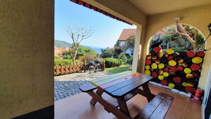 An outdoor patio with a wooden table and benches, a tiled floor, and a view of a garden and buildings in the distance. - German Village Gelbe Haus (Namhae)