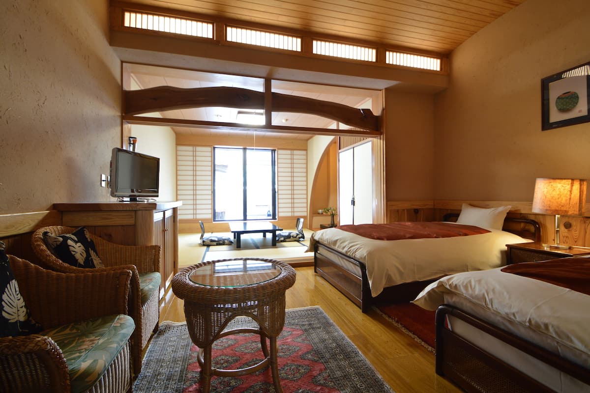Ein traditioneller japanischer Raum mit Tatami-Bodenbelag, niedriger Holzzieldecke und einer Schiebetür, die auf einen Balkon führt. Es gibt zwei Betten, einen Fernseher und einen kleinen Tisch mit einem Stuhl.