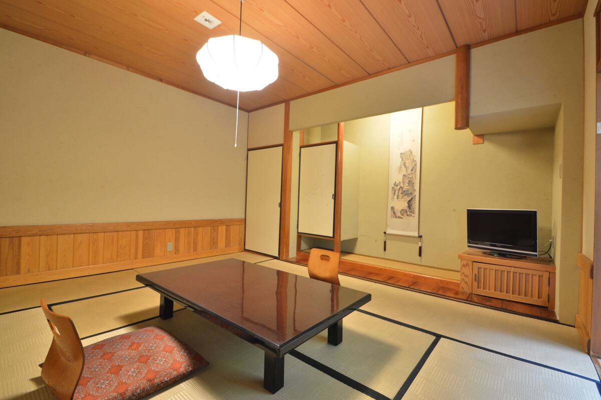 Ein Raum mit Tatami-Matten, einem niedrigen Tisch, Kissen, einem Fernseher auf einem Holzständer und einem traditionellen japanischen Gemälde an der Wand.