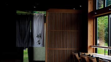 A dimly lit room with a wooden table and chairs, a large window, and a sliding door with a curtain.