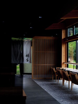 A dimly lit room with a wooden table and chairs, a large window, and a sliding door with a curtain. - Kurokawa Onsen Okunoyu (Minamioguni)