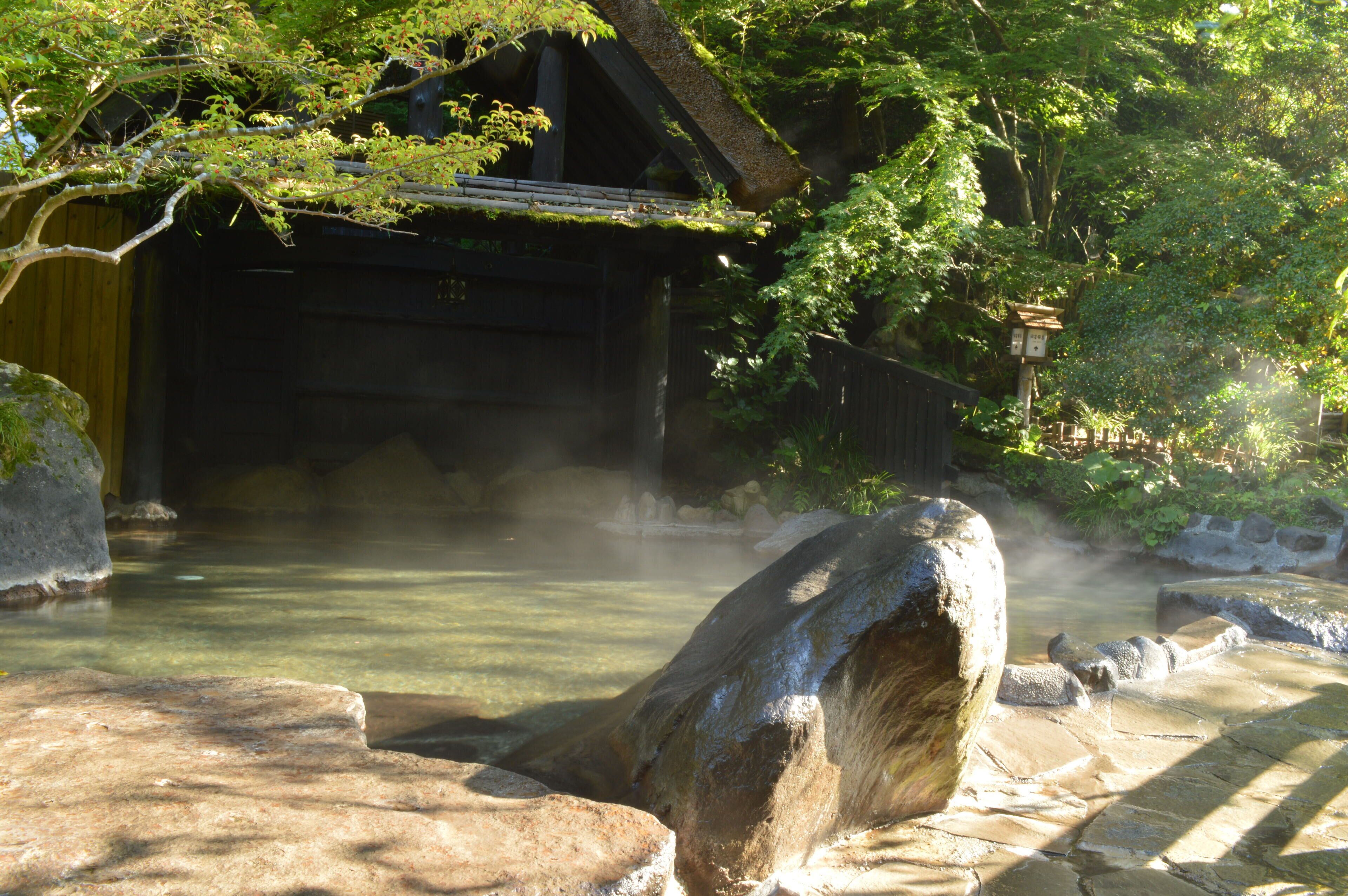 A traditional Japanese onsen with a wooden structure, a stone bath, and a large rock in the foreground.