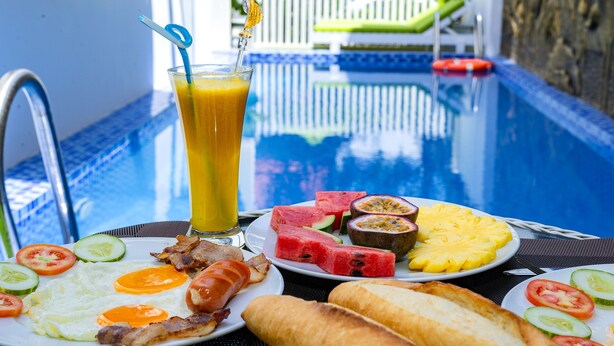A breakfast setting with a poolside table, including a glass of orange juice, a plate of eggs, bacon, toast, and various fruits. - Green Hill Hotel (Da Nang)
