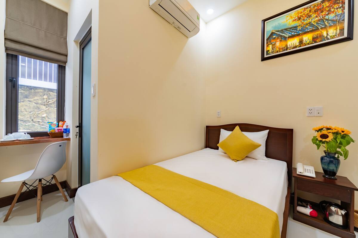 Quarto de hotel com cama, escrivaninha, cadeira, janela, ar condicionado, um quadro e um vaso com flores.