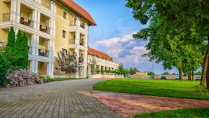 A multi-story hotel with a brick pathway leading to the entrance, surrounded by greenery and a clear sky. - Arcanum Hotel (Bekescsaba)