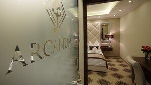 A hotel room with a bed, a nightstand, a lamp, and a view of the adjacent room. - Arcanum Hotel (Bekescsaba)