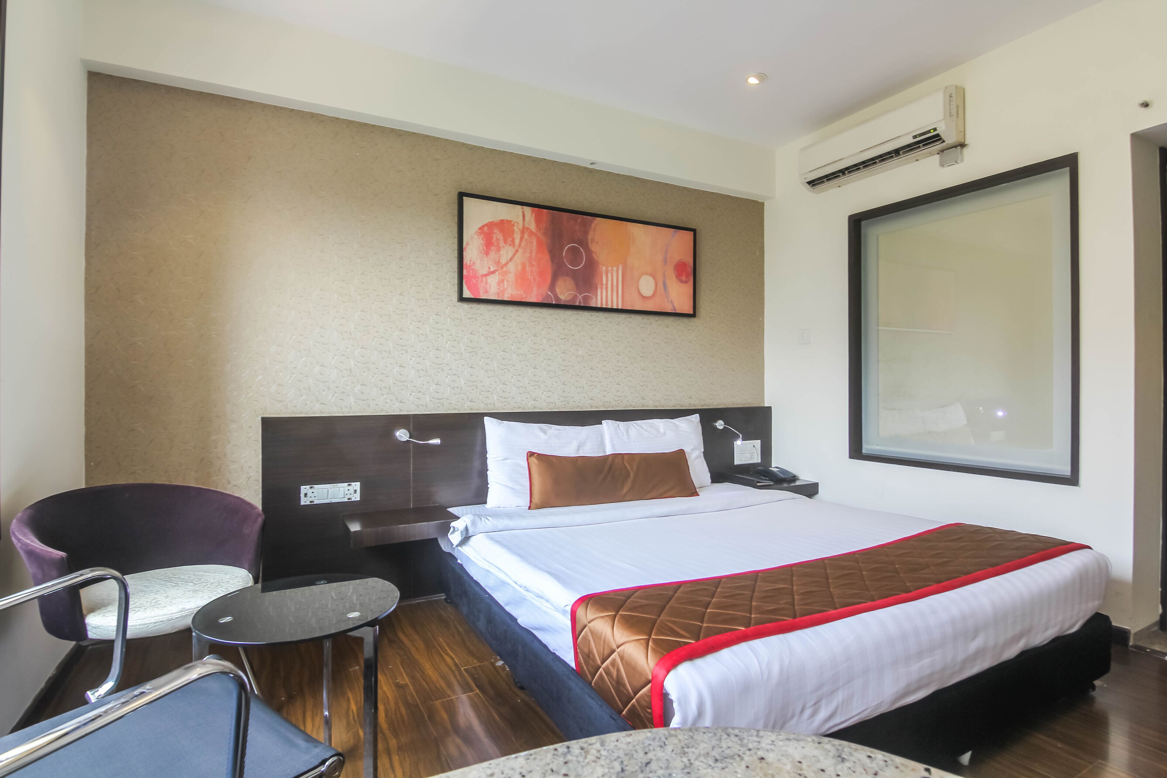 A hotel room with a bed, a chair, a small table, and a wall-mounted air conditioner.