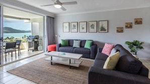 A modern living room with a grey sofa, a white coffee table, and a red chair. There are framed pictures on the wall and a plant on the right. - Poinciana Lodge 111, Seaview Apartment on Hamilton Island (Hamilton Island)