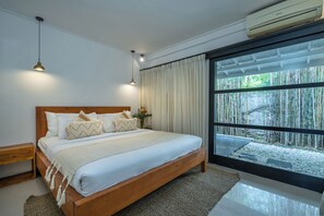 Une chambre avec un lit en bois, du linge de lit blanc, une table de chevet, une lampe et une grande fenêtre donnant sur un jardin de bambous.