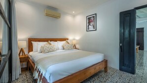A hotel room with a wooden bed, white bedding, a wall-mounted air conditioner, a framed photo, and patterned floor tiles. - 10-bedroom private estate in Seminyak, perfect for large groups, family gatherings, retreats, and celebrations. Enjoy daily breakfast, a private chef, airport transfers, complimentary massages, housekeeping, and a dedicated driver. (Seminyak)