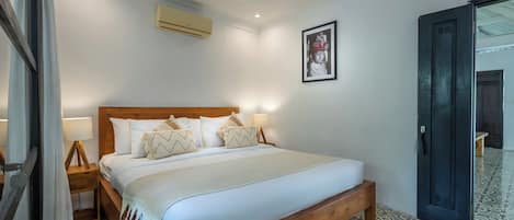 Habitación de hotel con cama de madera, ropa de cama blanca, aire acondicionado de pared, una fotografía enmarco y baldosas de suelo estampadas.