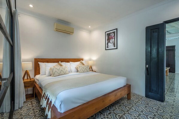 A hotel room with a wooden bed, white bedding, a wall-mounted air conditioner, a framed photo, and patterned floor tiles.