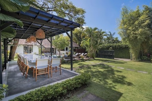 A covered outdoor dining area with a table and chairs, surrounded by greenery and a clear sky. - 10-bedroom private estate in Seminyak, perfect for large groups, family gatherings, retreats, and celebrations. Enjoy daily breakfast, a private chef, airport transfers, complimentary massages, housekeeping, and a dedicated driver. (Seminyak)