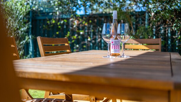 A wooden table with a bottle of wine and two glasses, set outdoors with a wooden chair and a green fence in the background. - Flinders Ranges Bed and Breakfast (HAWKER)