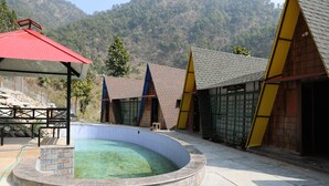 A swimming pool with a stone edge, surrounded by buildings with colorful roofs and a mountainous backdrop. - Rakatal Rocks Resort (Lansdowne)