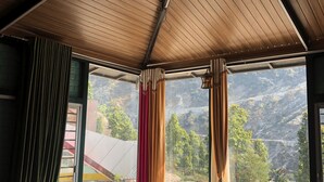 A modern room with a glass coffee table, a sofa, and a view of a mountainous landscape through large windows. - Rakatal Rocks Resort (Lansdowne)