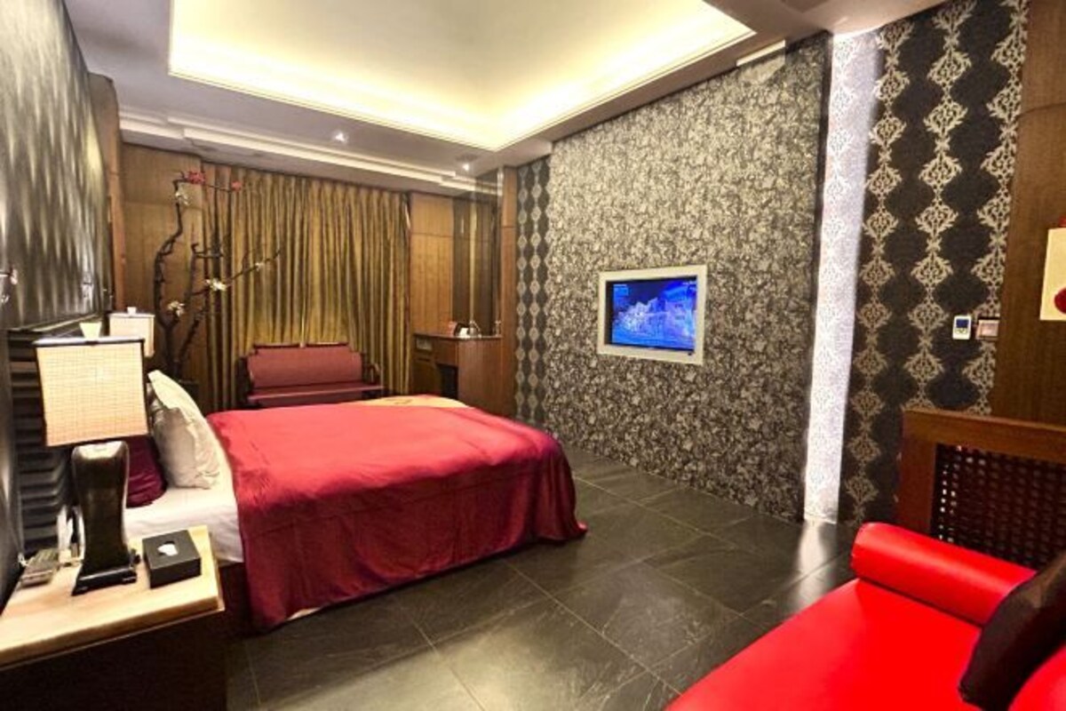 A hotel room with a bed, a desk with a lamp, a chair, and a television on the wall.