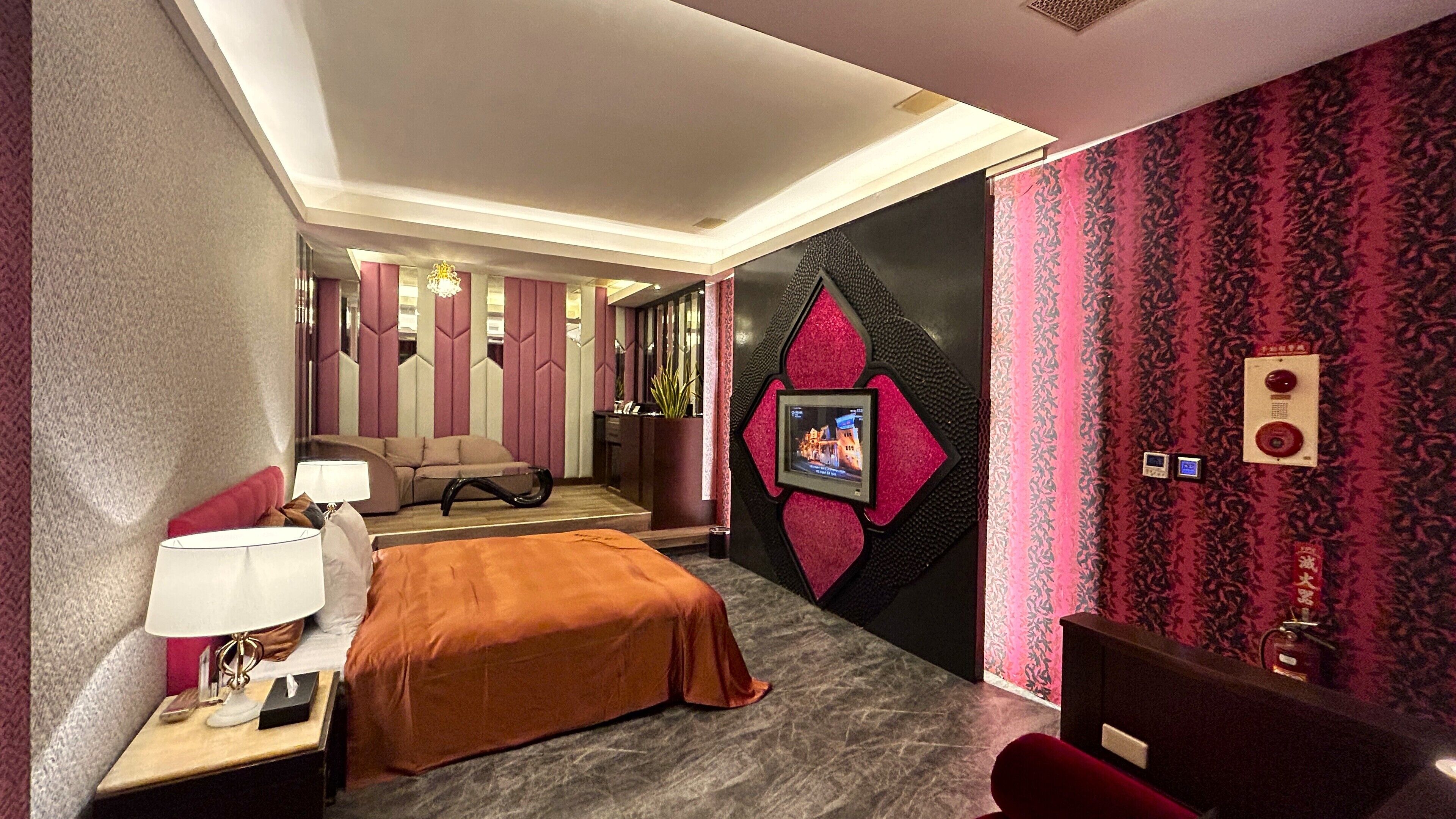Habitación de hotel con cama, escritorio con lámpara, televisor montado en la pared y una silla roja.