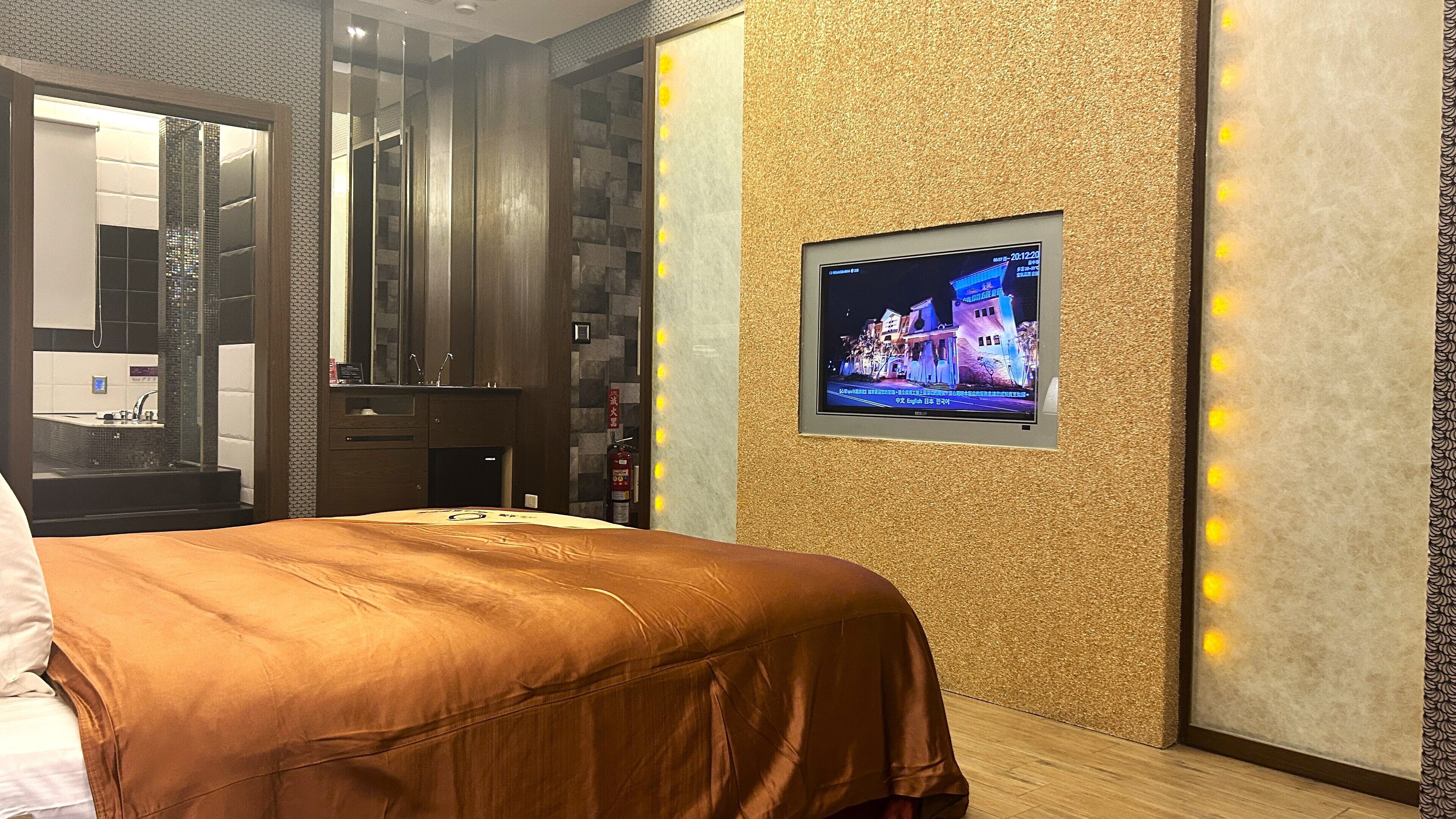 A hotel room with a bed, a television mounted on the wall, and a bathroom visible through a glass partition.