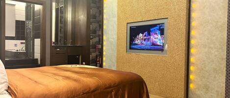 A hotel room with a bed, a television mounted on the wall, and a bathroom visible through a glass partition.