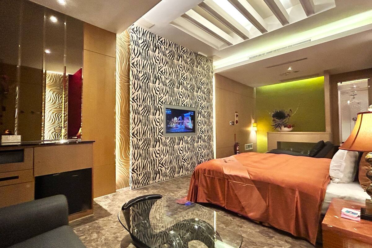 A hotel room with a zebra-patterned accent wall, a television, a bed with an orange bedspread, a desk with a chair, and a lamp.
