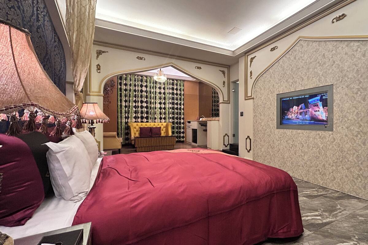 A hotel room with a large bed, a television on the wall, a small desk, and a decorative mirror.