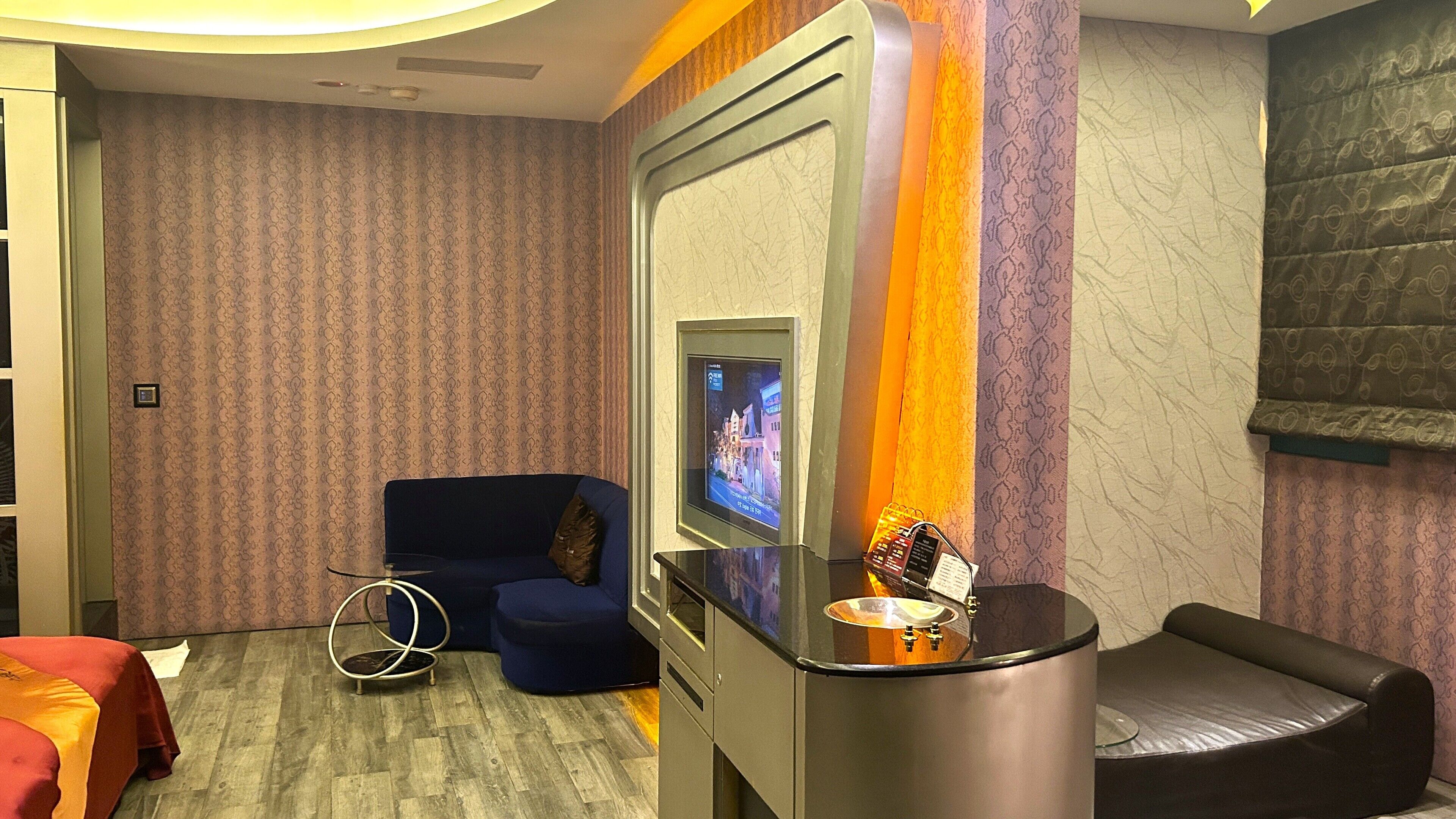Habitación de hotel con un sofá azul, una pequeña mesa redonda con una lámpara, un televisor montado en la pared y un papel tapiz estampado.