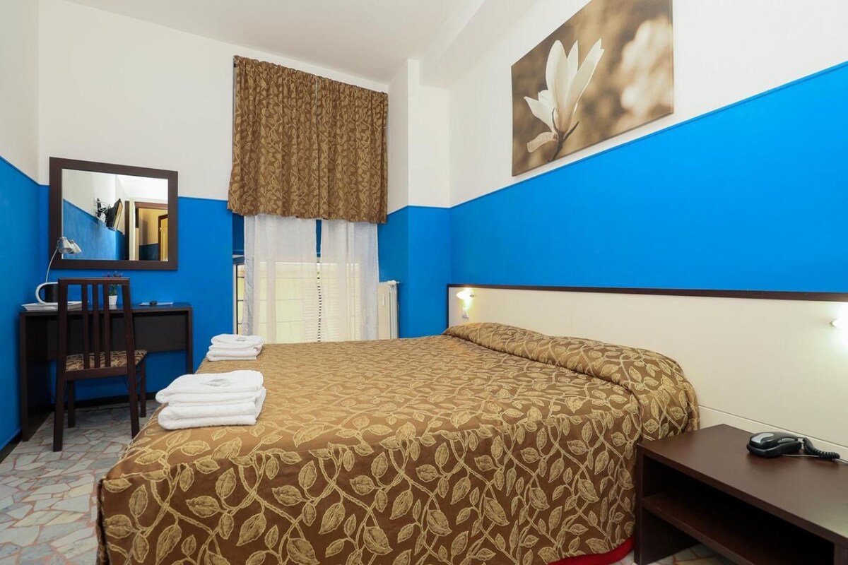 A hotel room with a bed, a desk, a chair, a mirror, and a telephone.